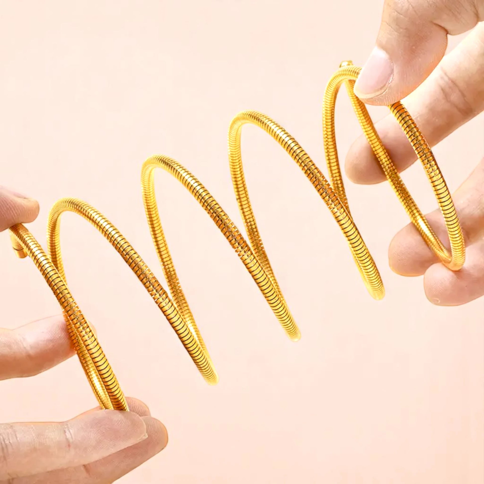 Two hands holding a swirling, shiny gold metal spring, showcasing its coiled design against a soft pink background.