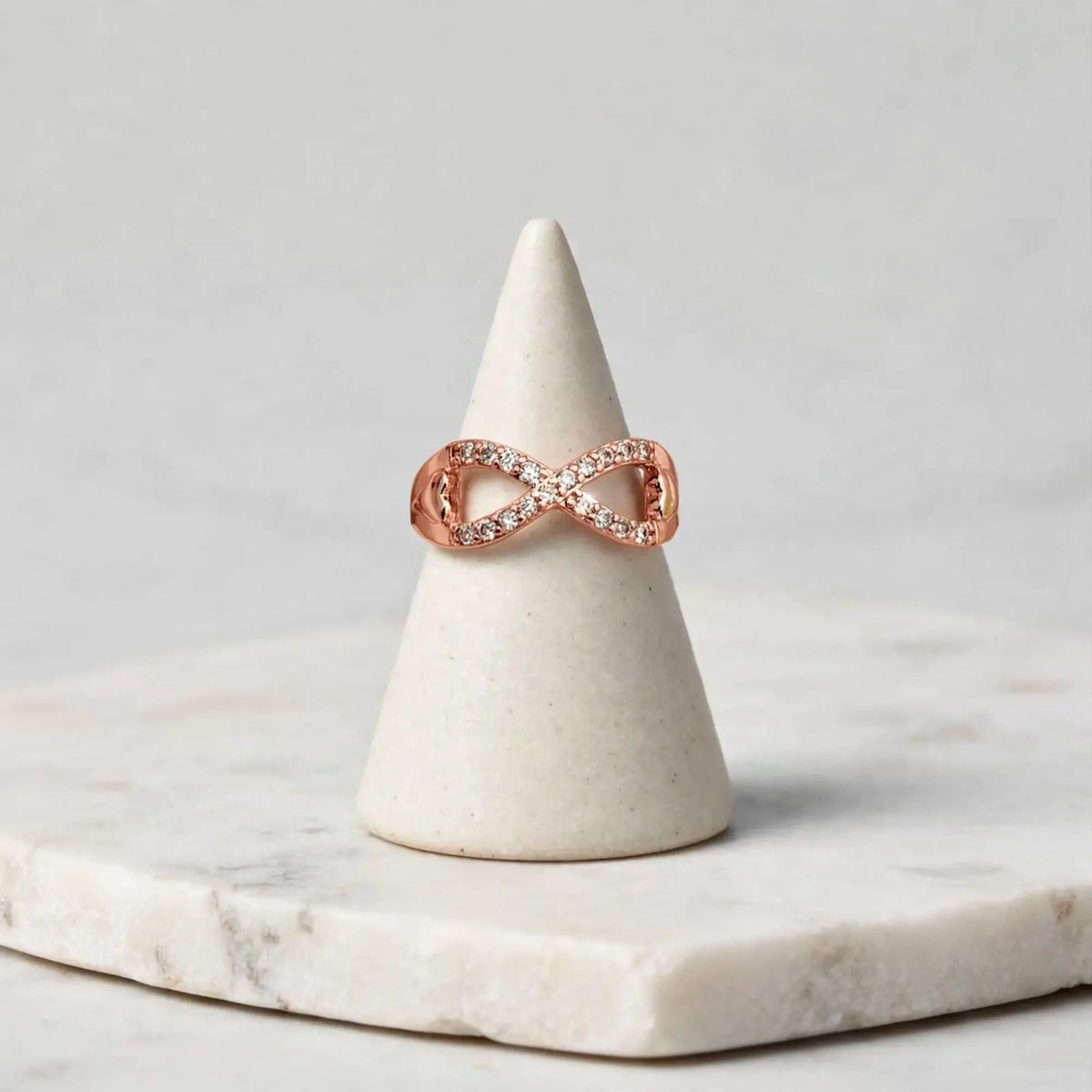 A rose gold infinity ring with small clear pavé stones, displayed on a white ceramic cone stand over a white marble slab against a light gray background.