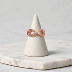 A rose gold infinity ring with small clear pavé stones, displayed on a white ceramic cone stand over a white marble slab against a light gray background.