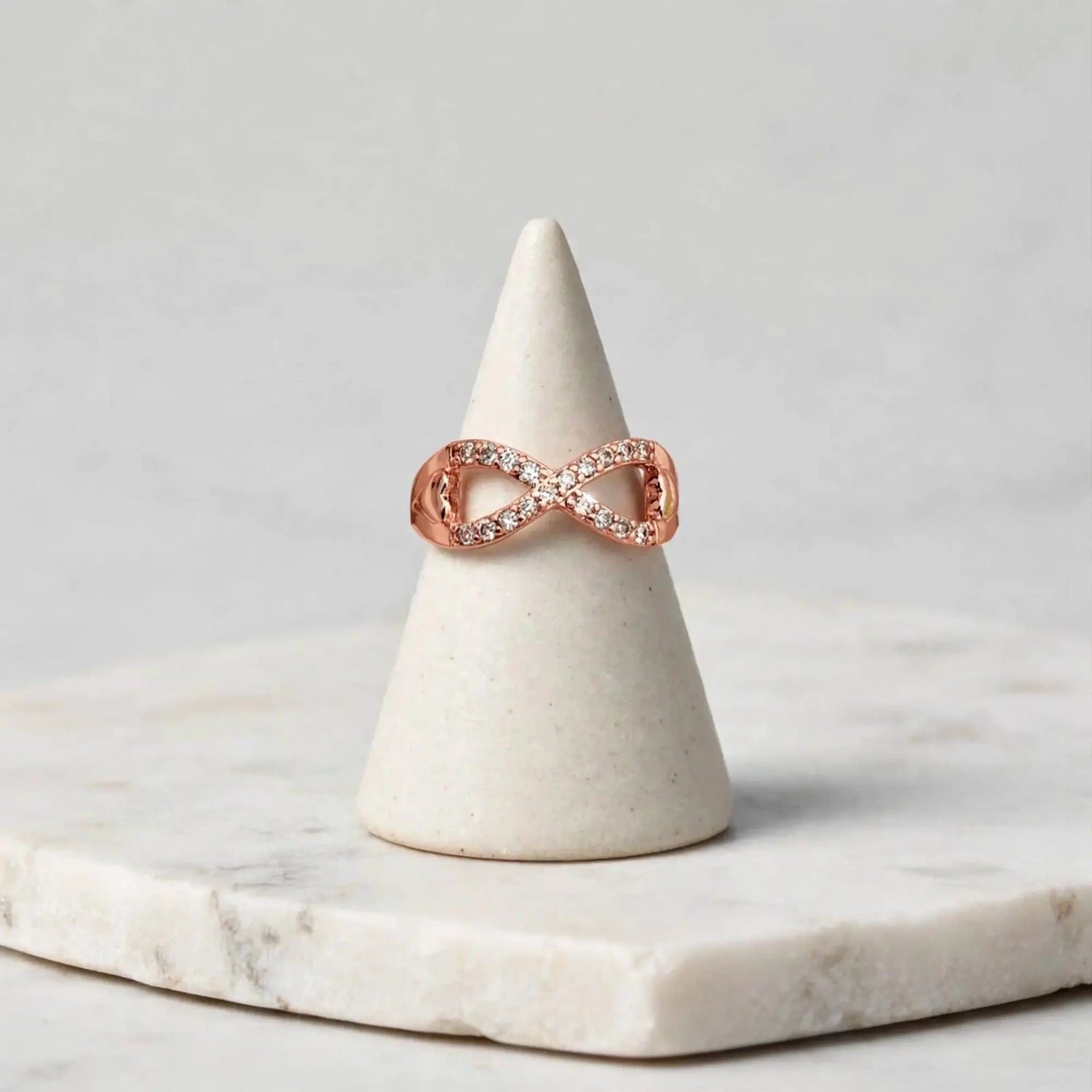 A rose gold infinity ring with small clear pavé stones, displayed on a white ceramic cone stand over a white marble slab against a light gray background.