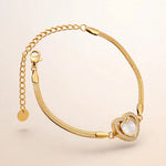 A gold snake chain bracelet with a heart-shaped charm featuring a white mother-of-pearl center and a border of small clear stones, shown on a light beige background.