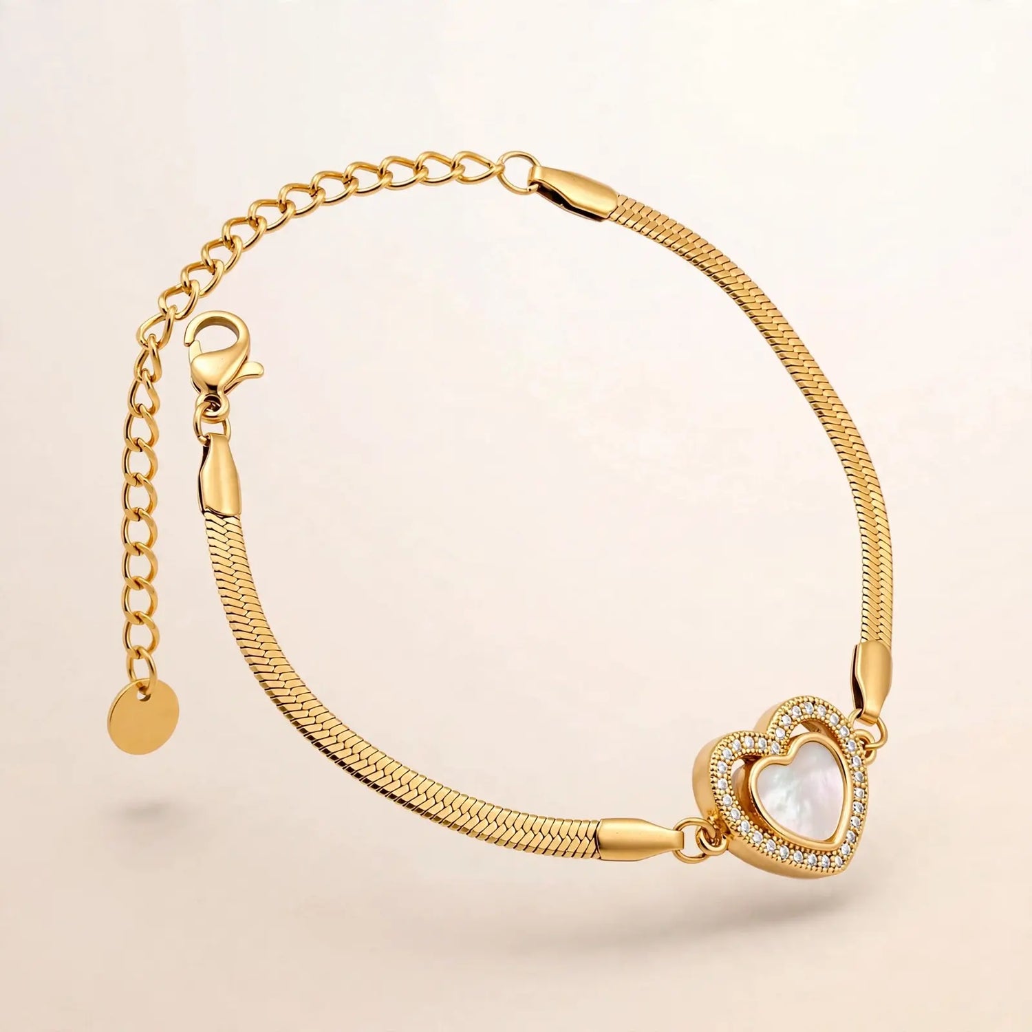 A gold snake chain bracelet with a heart-shaped charm featuring a white mother-of-pearl center and a border of small clear stones, shown on a light beige background.