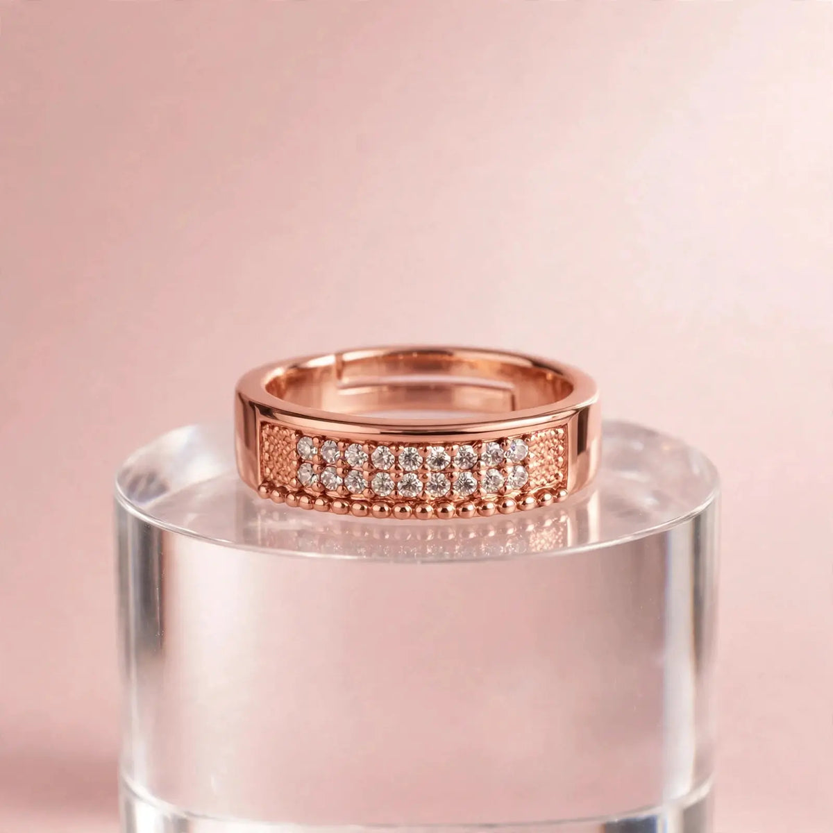 A rose gold ring with a row of clear pavé-set stones and textured side panels, displayed on a clear acrylic pedestal against a soft pink background.