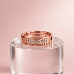 A rose gold ring with a row of clear pavé-set stones and textured side panels, displayed on a clear acrylic pedestal against a soft pink background.
