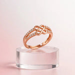 A rose gold ring with a central infinity symbol and pavé-set clear stones, displayed on a clear acrylic stand against a soft pink background.