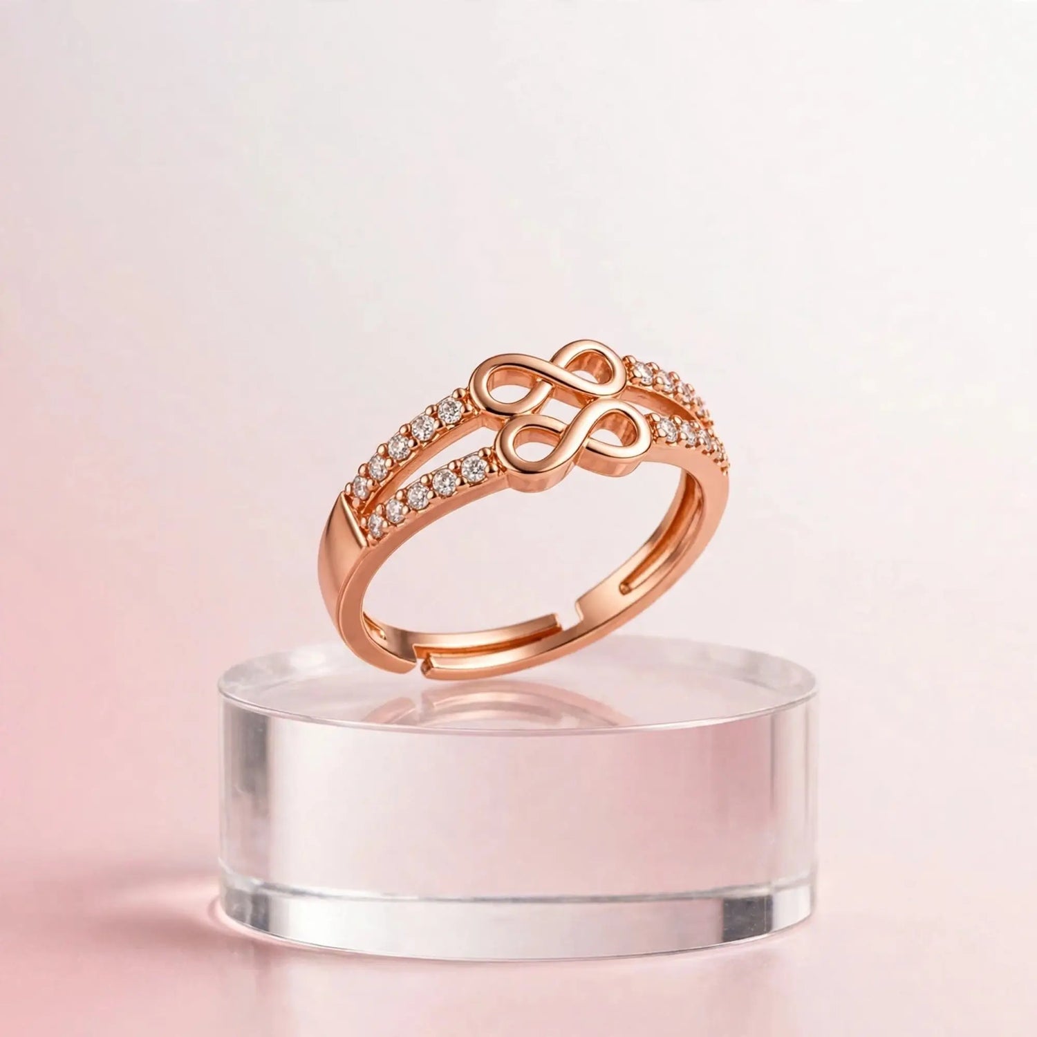 A rose gold ring with a central infinity symbol and pavé-set clear stones, displayed on a clear acrylic stand against a soft pink background.