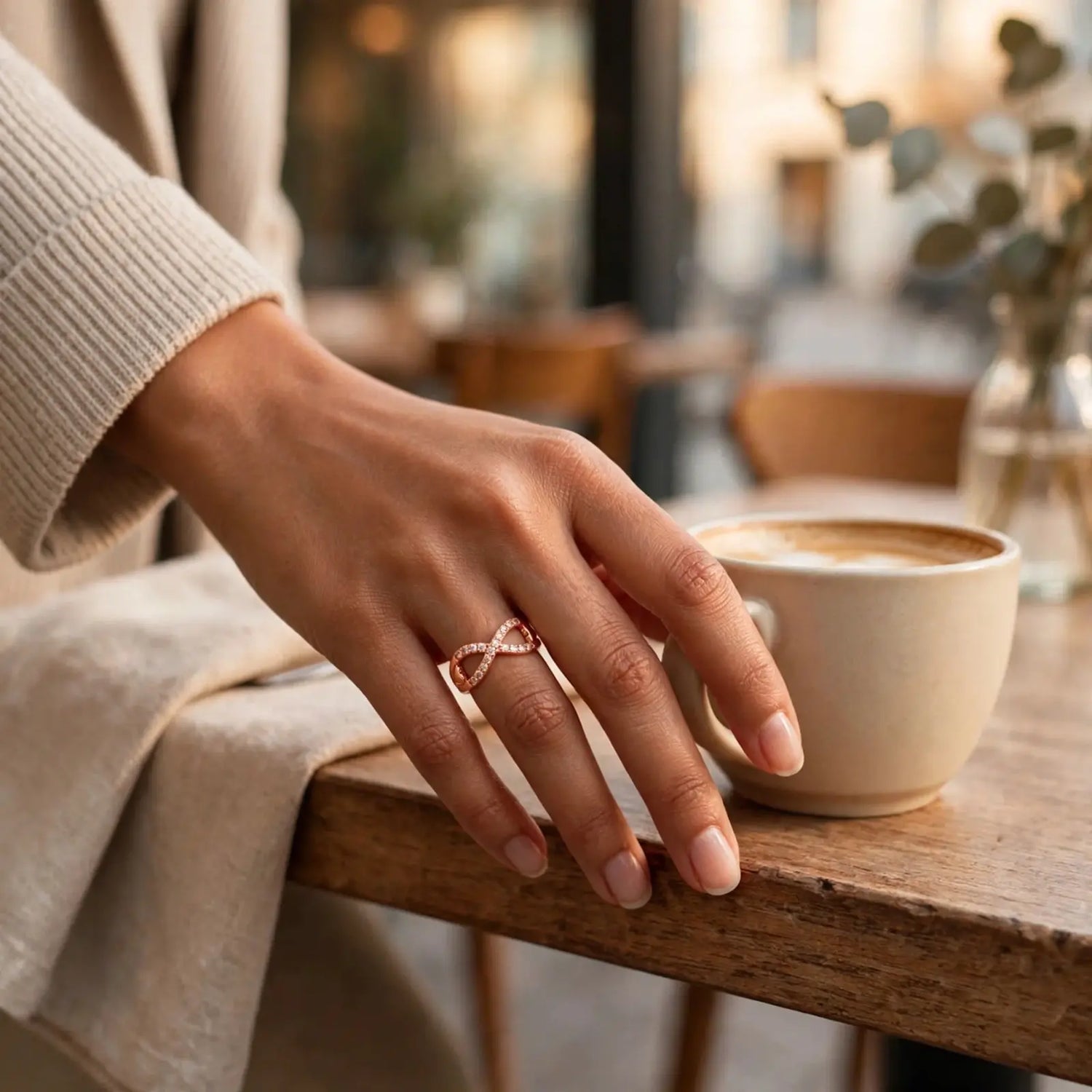 A close-up lifestyle shot of a person's hand resting on a wooden table next to a ceramic coffee cup, wearing a delicate rose gold infinity ring with small clear stones.