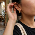 A close-up lifestyle shot of a person wearing a small, textured gold hoop earring. The person is wearing a black ribbed top and a beige canvas shoulder strap, with a softly blurred cafe interior in the background.