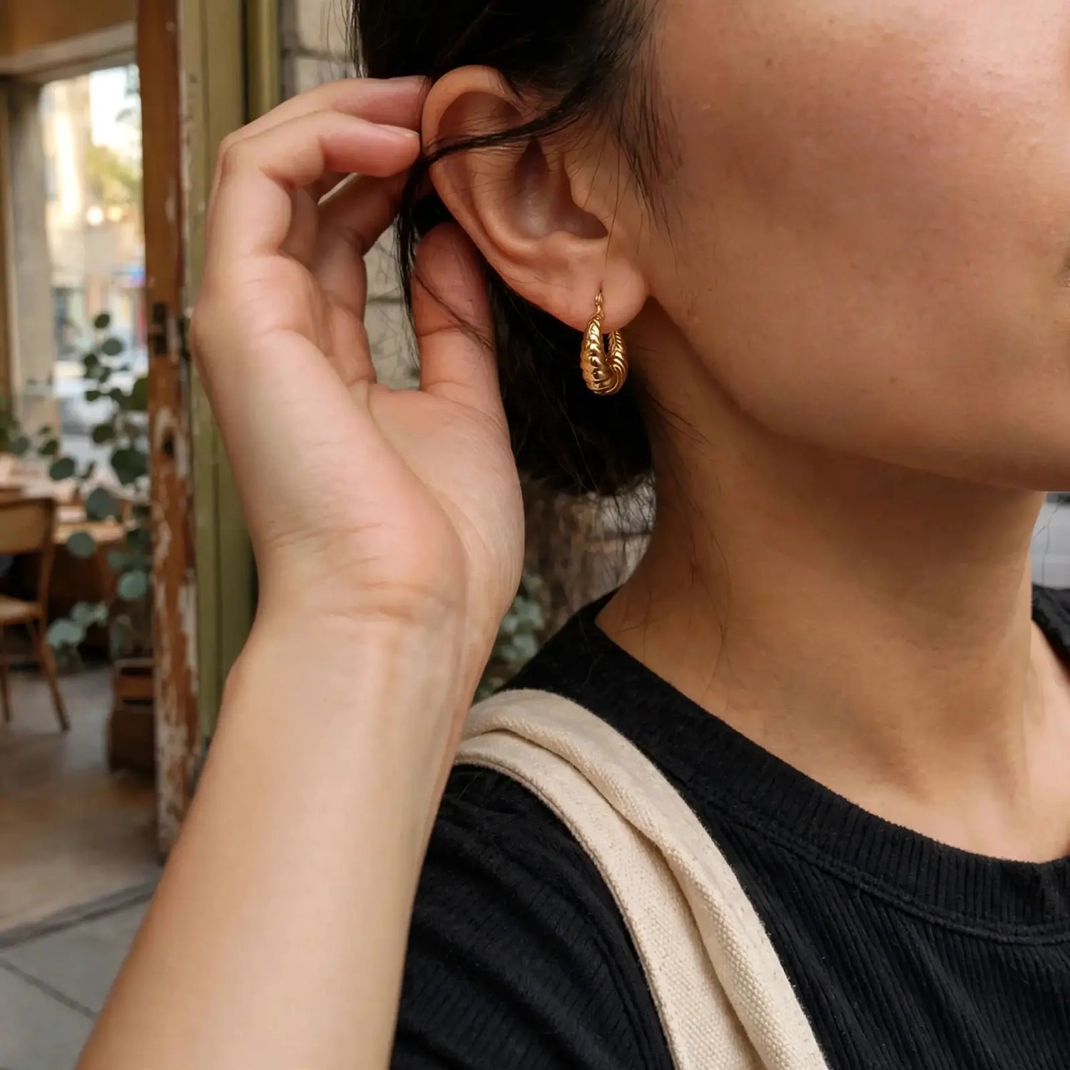 A close-up lifestyle shot of a person wearing a small, textured gold hoop earring. The person is wearing a black ribbed top and a beige canvas shoulder strap, with a softly blurred cafe interior in the background.
