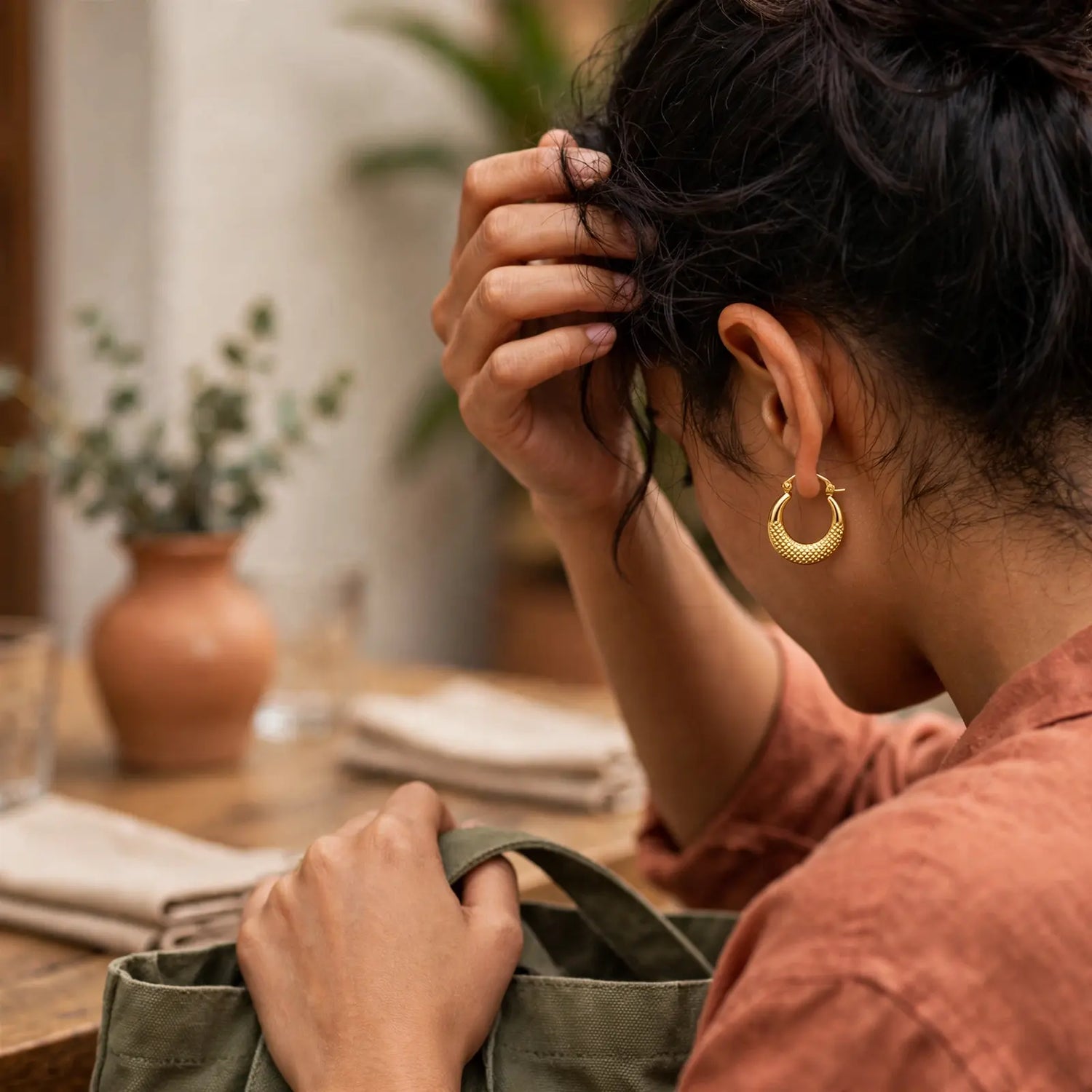 A woman in a terracotta shirt wearing a textured gold huggie hoop earring, shown from a side-back angle in a warm, softly lit indoor setting.