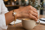 A gold heart-shaped charm bracelet with a white inlay and a small dangling charm, worn on a person's wrist while they stir a cup of coffee in a cafe setting.