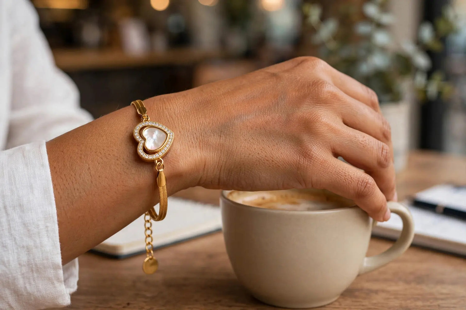 A gold heart-shaped charm bracelet with a white inlay and a small dangling charm, worn on a person's wrist while they stir a cup of coffee in a cafe setting.