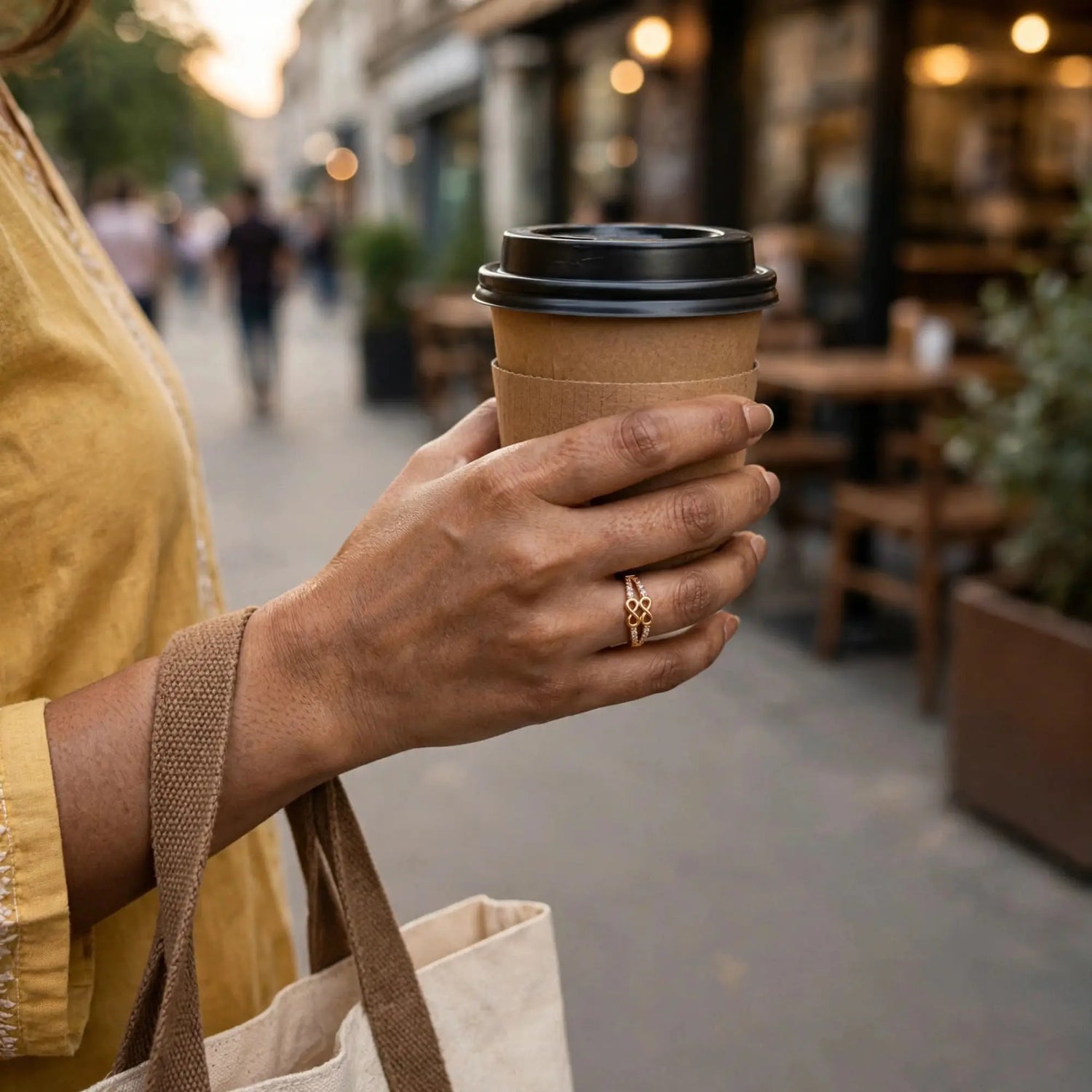 A person in a mustard yellow top holds a brown paper coffee cup with a black lid on a blurred city sidewalk. The person is wearing a gold ring and carrying a beige tote bag.