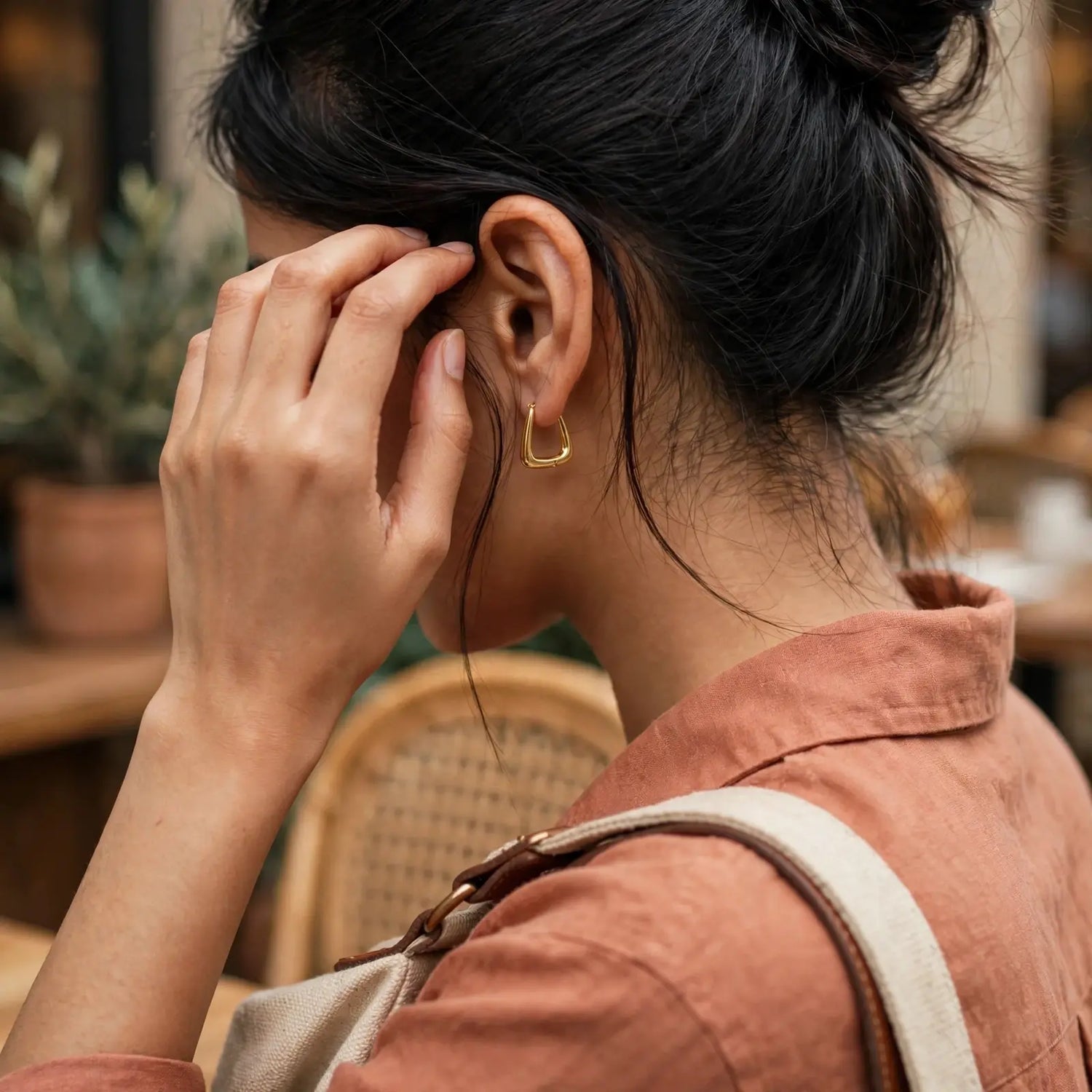 A woman in profile wearing a small gold hoop earring, with her hand near her face in a softly blurred indoor setting.