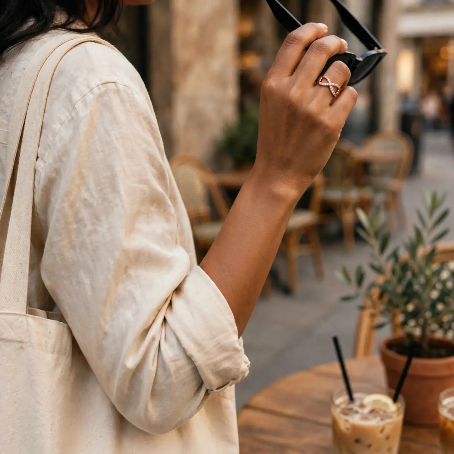 A person at an outdoor cafe wearing a delicate infinity-shaped ring with a rose gold-toned metal and clear gemstone, holding black sunglasses. The person is wearing a beige linen shirt and a canvas tote bag.