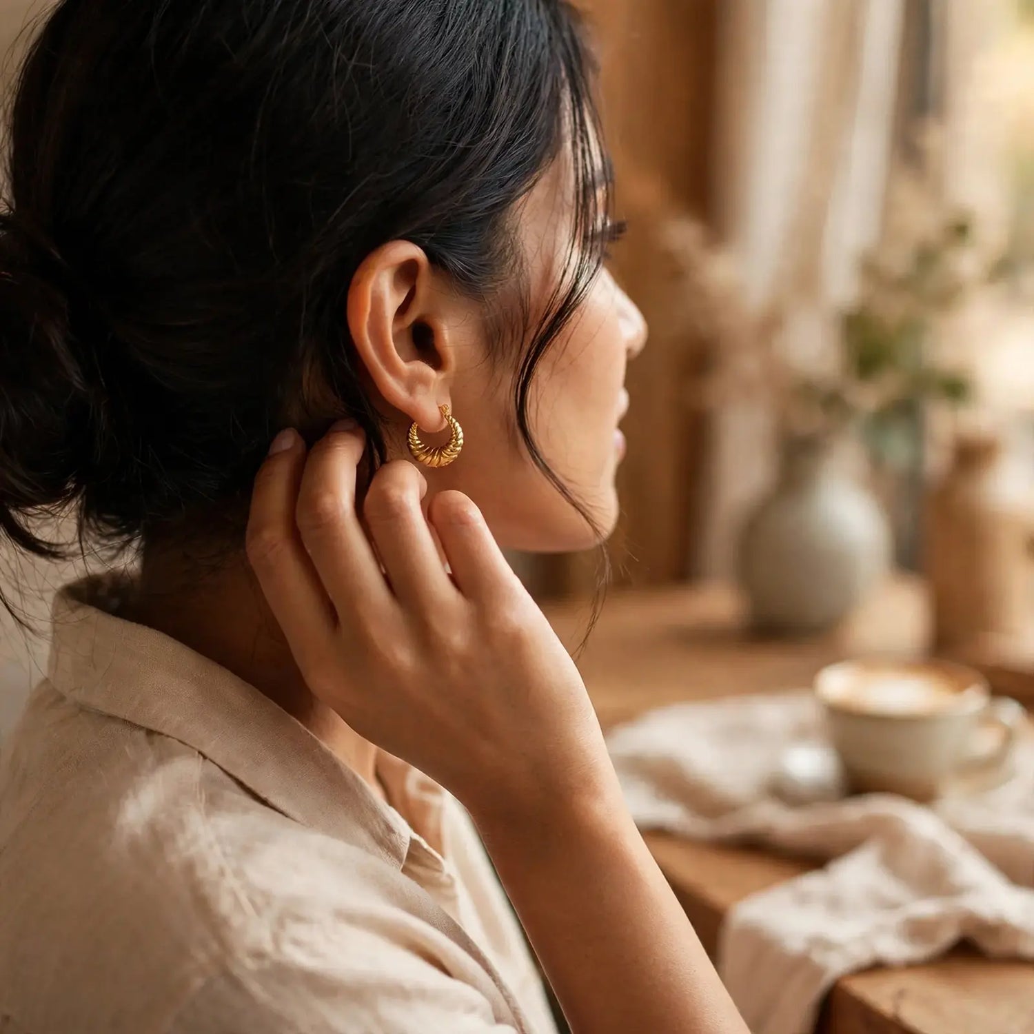 A woman in profile wearing a small gold hoop earring, shown in a warm, softly lit indoor setting with a wooden table and blurred decor in the background.