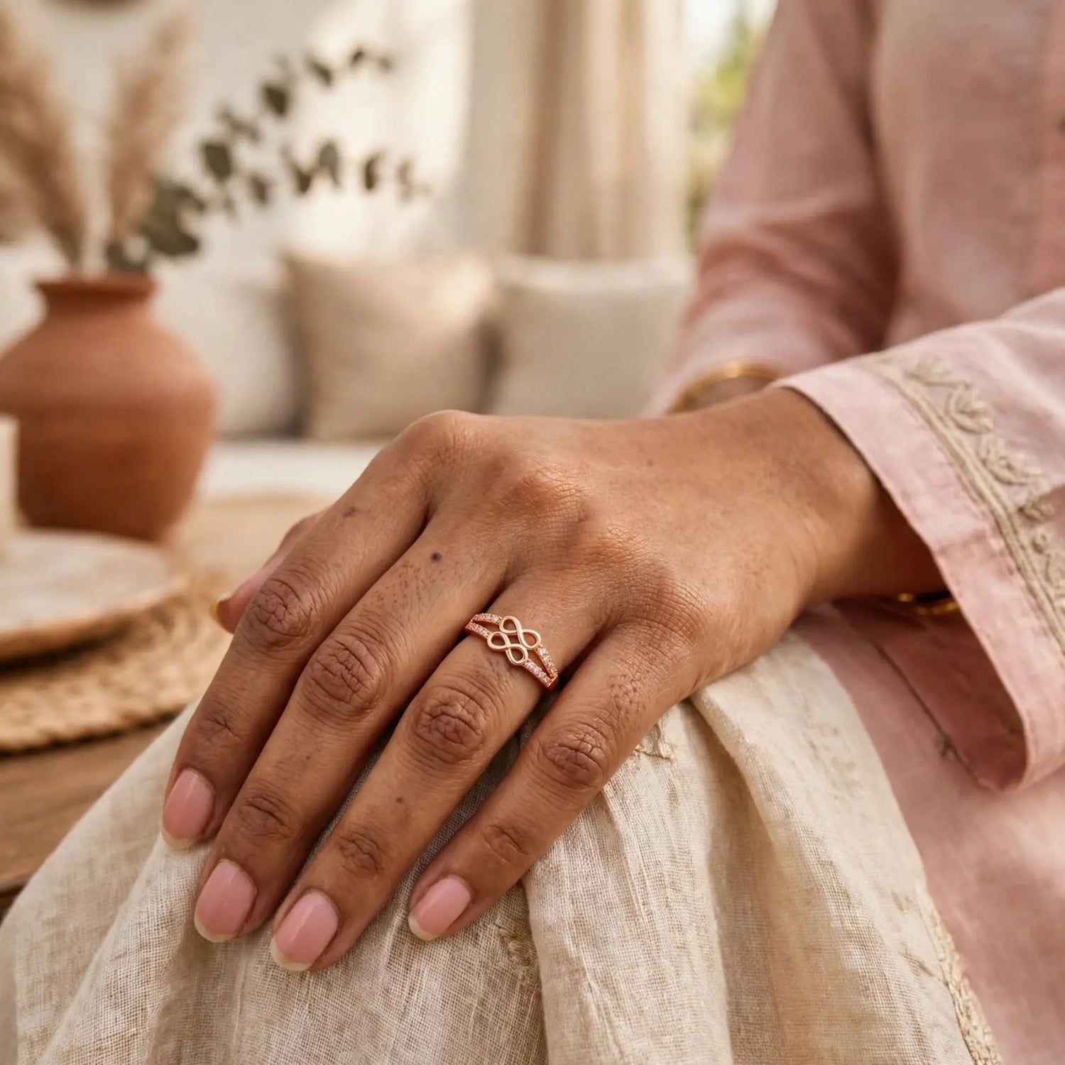 A close-up of a person's hand resting on cream-colored fabric, wearing a delicate rose gold ring with an intricate knot design. The background features a soft-focus terracotta vase and neutral-toned cushions.