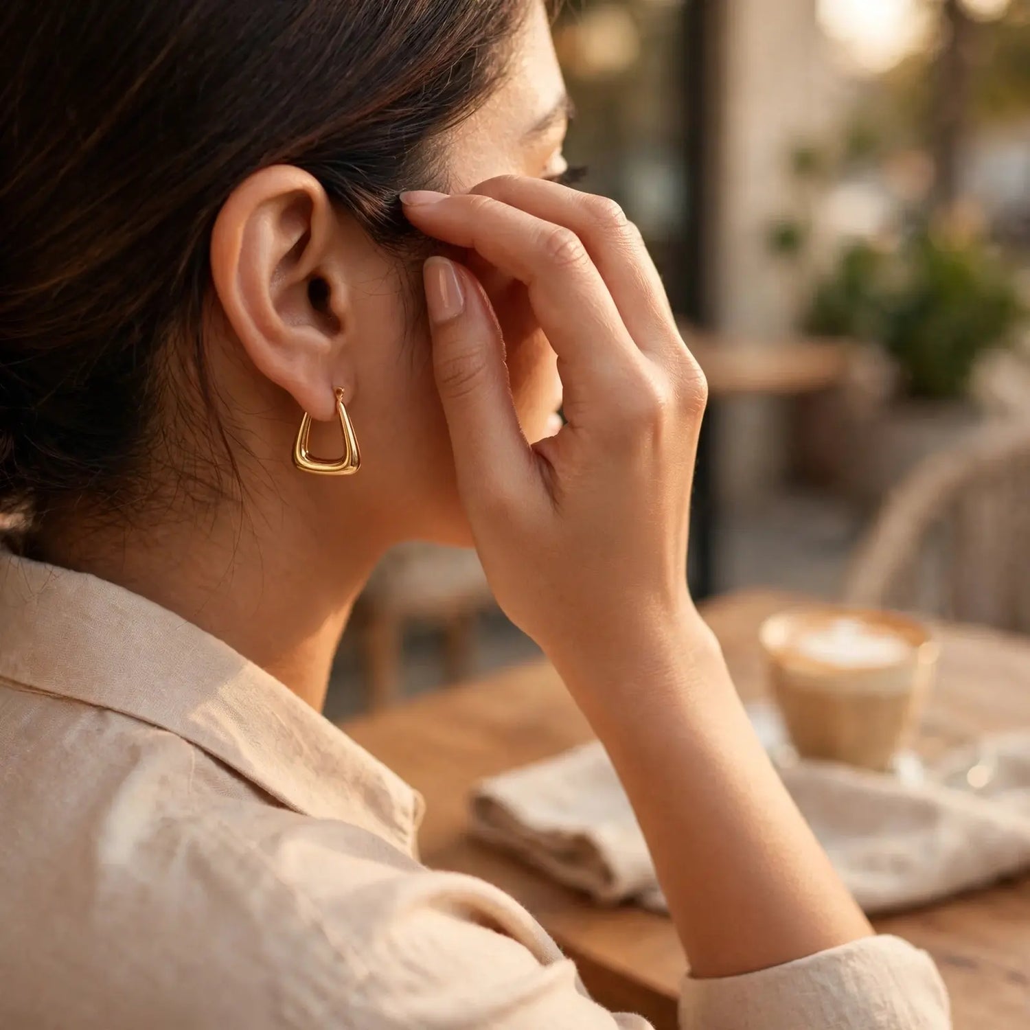 A woman in a beige shirt wearing a small gold triangular hoop earring, shown in profile in a warm cafe setting.