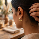 A close-up side profile of a woman wearing a textured gold crescent hoop earring, with her hand in her hair, set against a warm, softly blurred background.
