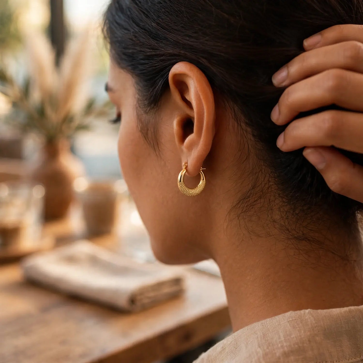 A close-up side profile of a woman wearing a textured gold crescent hoop earring, with her hand in her hair, set against a warm, softly blurred background.