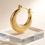 A polished gold hoop earring with a textured, woven-like pattern on the inner curve, displayed on a clear acrylic block against a warm beige background.