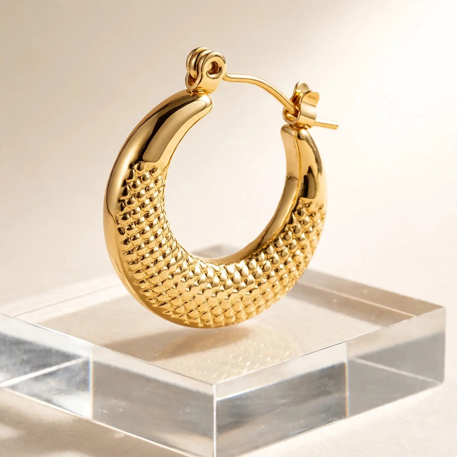 A polished gold hoop earring with a textured, woven-like pattern on the inner curve, displayed on a clear acrylic block against a warm beige background.
