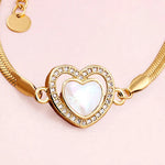 Gold-plated snake chain bracelet featuring a heart-shaped pendant with a white iridescent mother-of-pearl center and a border of clear rhinestones, set against a solid pale pink background.