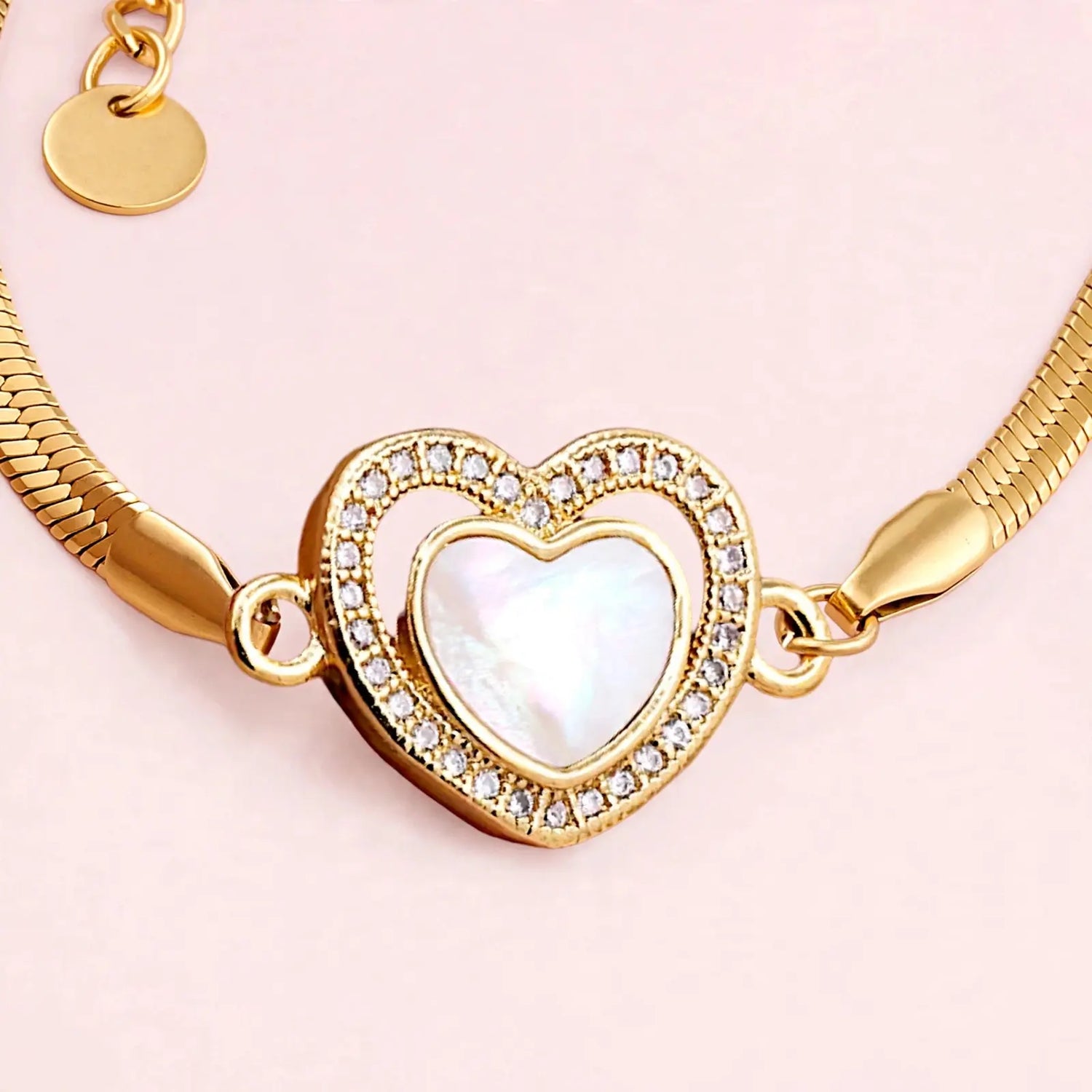 Gold-plated snake chain bracelet featuring a heart-shaped pendant with a white iridescent mother-of-pearl center and a border of clear rhinestones, set against a solid pale pink background.