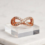 A rose gold infinity ring with pavé-set clear stones, displayed on a clear acrylic block over a white marble surface.
