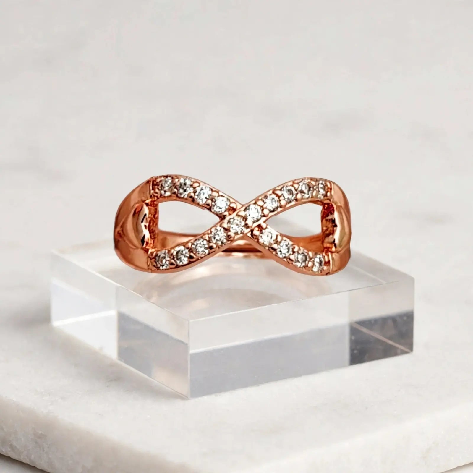 A rose gold infinity ring with pavé-set clear stones, displayed on a clear acrylic block over a white marble surface.