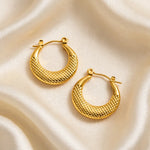 A pair of textured gold crescent hoop earrings displayed on a draped cream satin fabric background.
