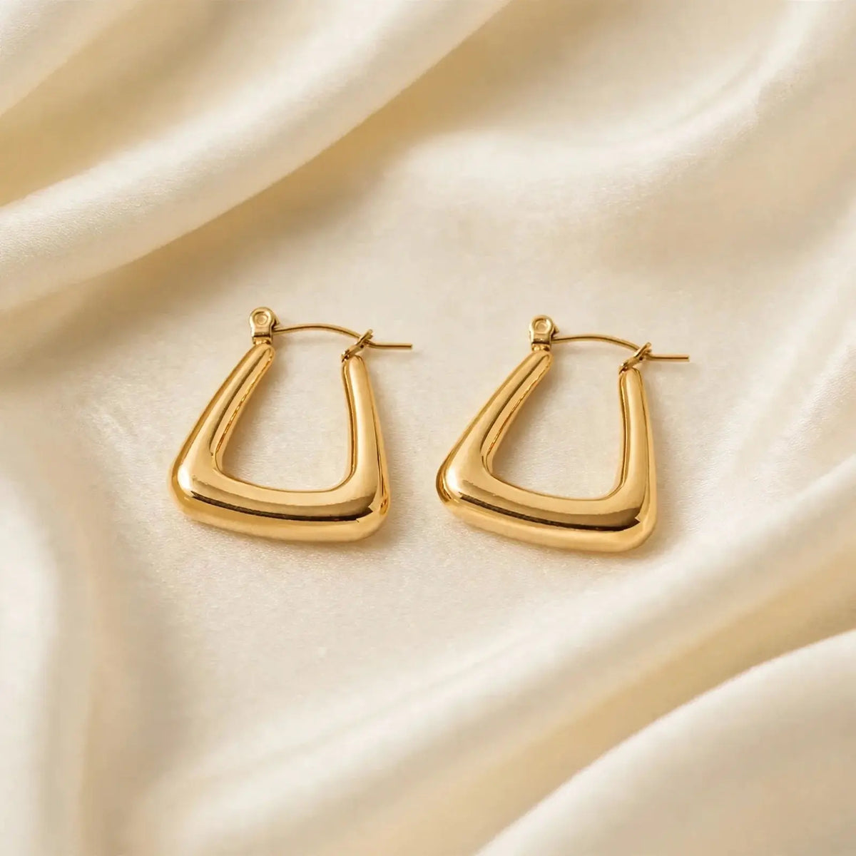 A pair of gold-toned, abstract open hoop earrings with a polished finish, displayed on a draped cream-colored satin fabric.