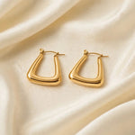 A pair of gold-toned, abstract open hoop earrings with a polished finish, displayed on a draped cream-colored satin fabric.