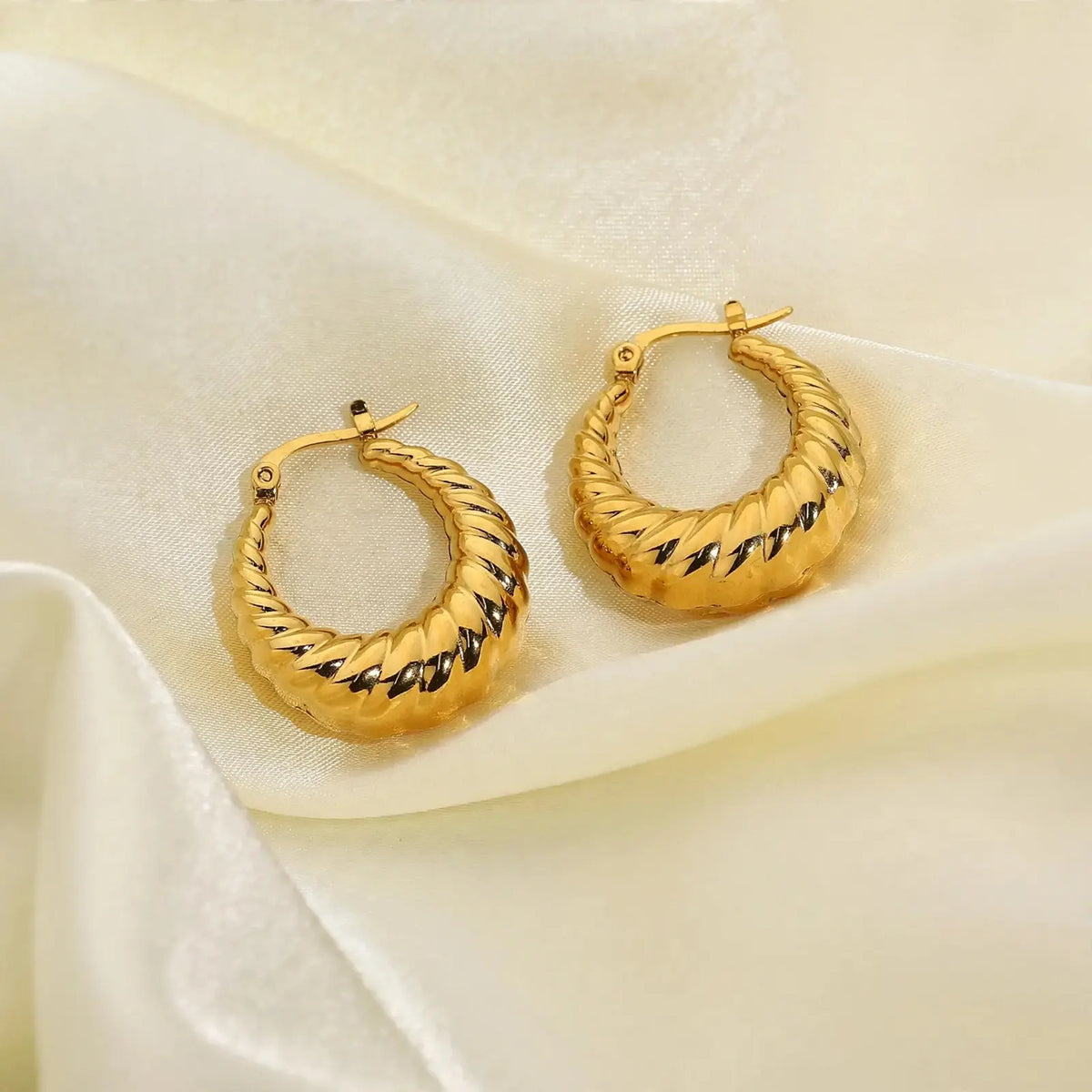 A pair of gold-toned twisted hoop earrings with black lightning-bolt accents, displayed on draped cream satin fabric.