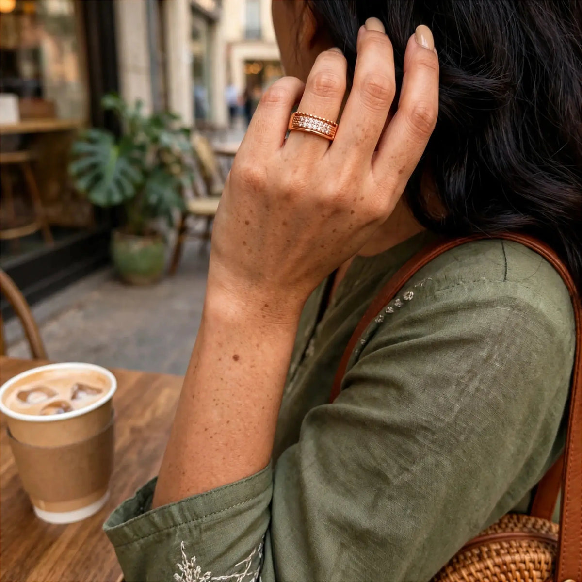 A close-up lifestyle shot of a woman wearing a rose gold band ring with pavé-set clear stones. She is sitting at a cafe table with a coffee cup nearby, wearing an olive green shirt and a brown woven shoulder bag.