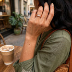 A close-up lifestyle shot of a woman wearing a rose gold band ring with pavé-set clear stones. She is sitting at a cafe table with a coffee cup nearby, wearing an olive green shirt and a brown woven shoulder bag.
