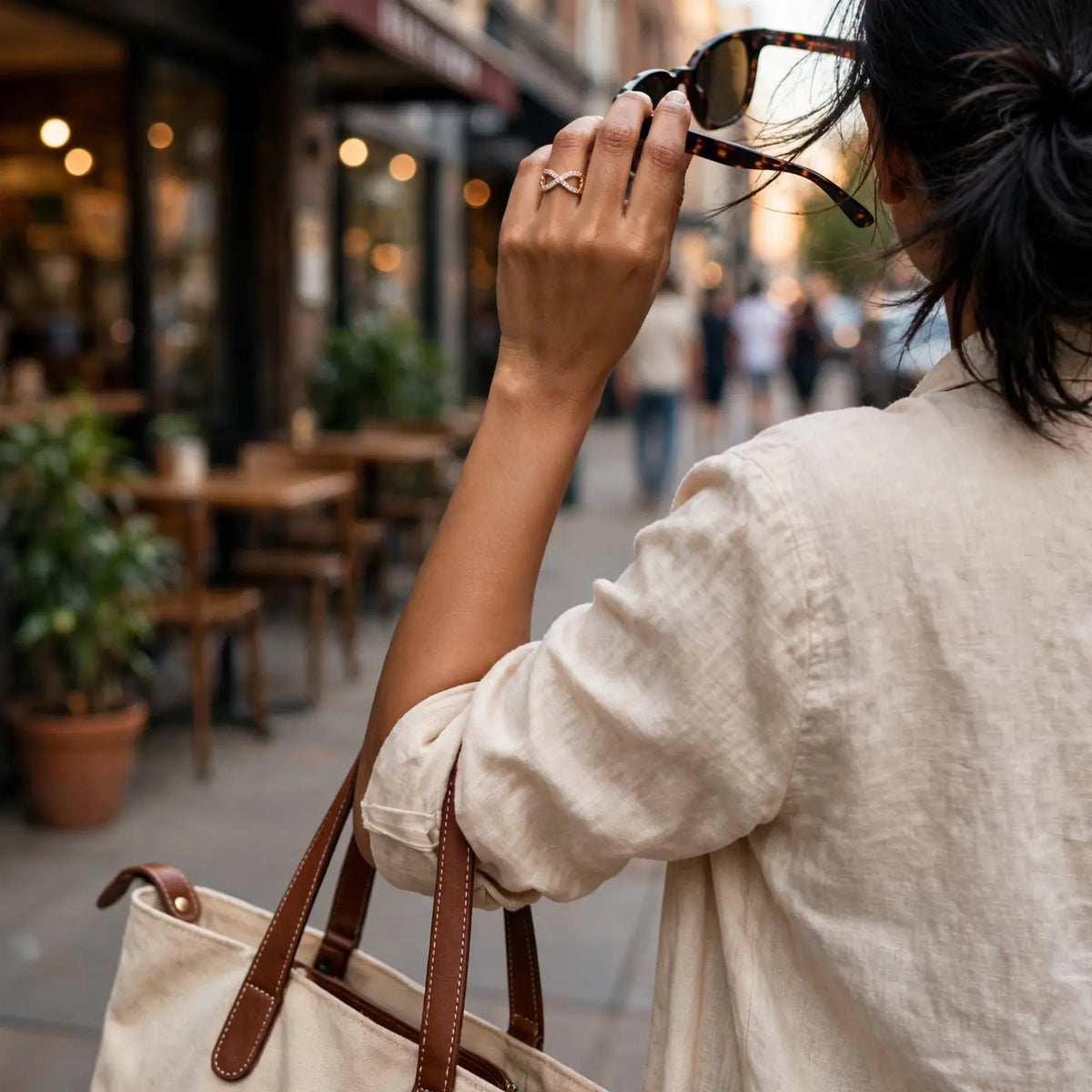 A woman in a light beige linen shirt carries a cream canvas tote bag with brown leather handles while walking on a city street, holding tortoiseshell sunglasses near her face.