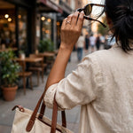 A woman in a light beige linen shirt carries a cream canvas tote bag with brown leather handles while walking on a city street, holding tortoiseshell sunglasses near her face.