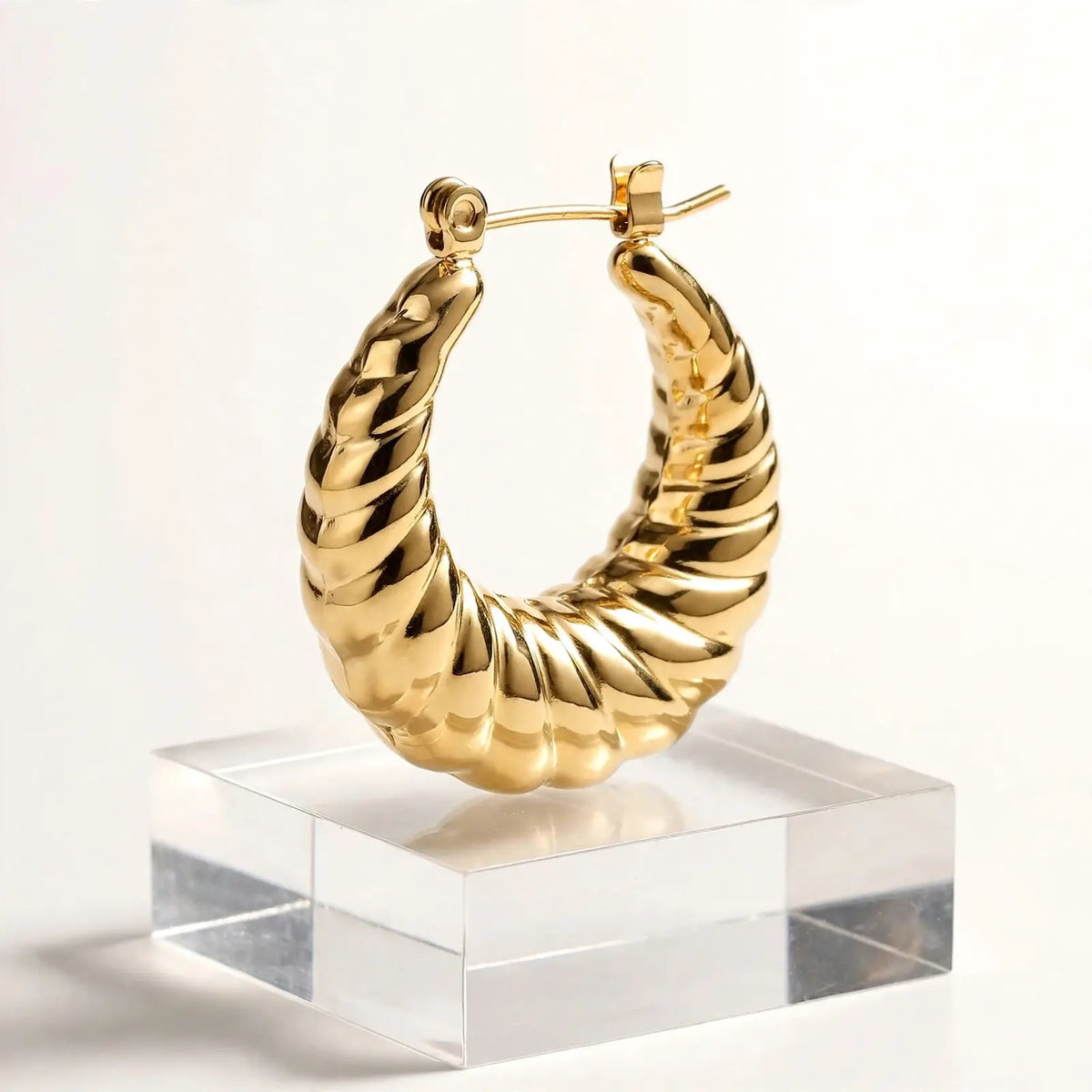 A gold hoop earring with a twisted, rope-like textured design, displayed on a clear acrylic block against a clean white background.