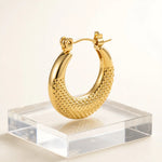 A polished gold crescent hoop earring with a textured, diamond-patterned lower half, displayed on a clear acrylic block against a clean white background.