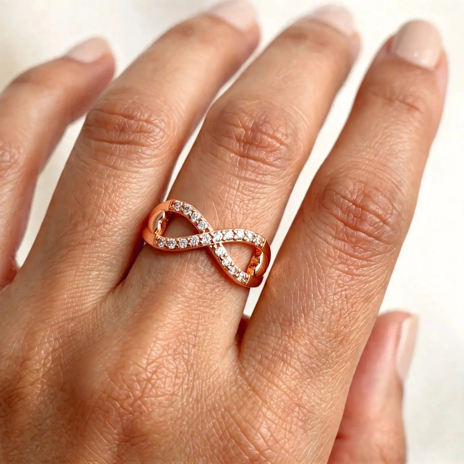 A close-up of a hand wearing a rose gold infinity ring set with small clear stones. The ring features an intertwined loop design, with one side paved with tiny clear gemstones and the other side featuring polished rose gold.
