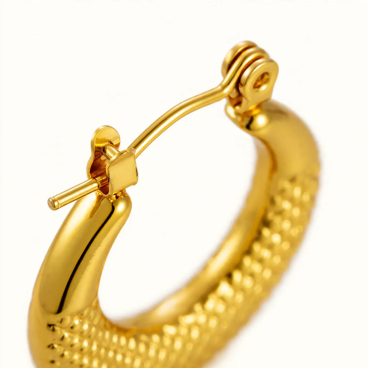 A close-up of a gold hoop earring with a textured, beaded outer band and a secure spring-ring clasp, shown on a plain white background.