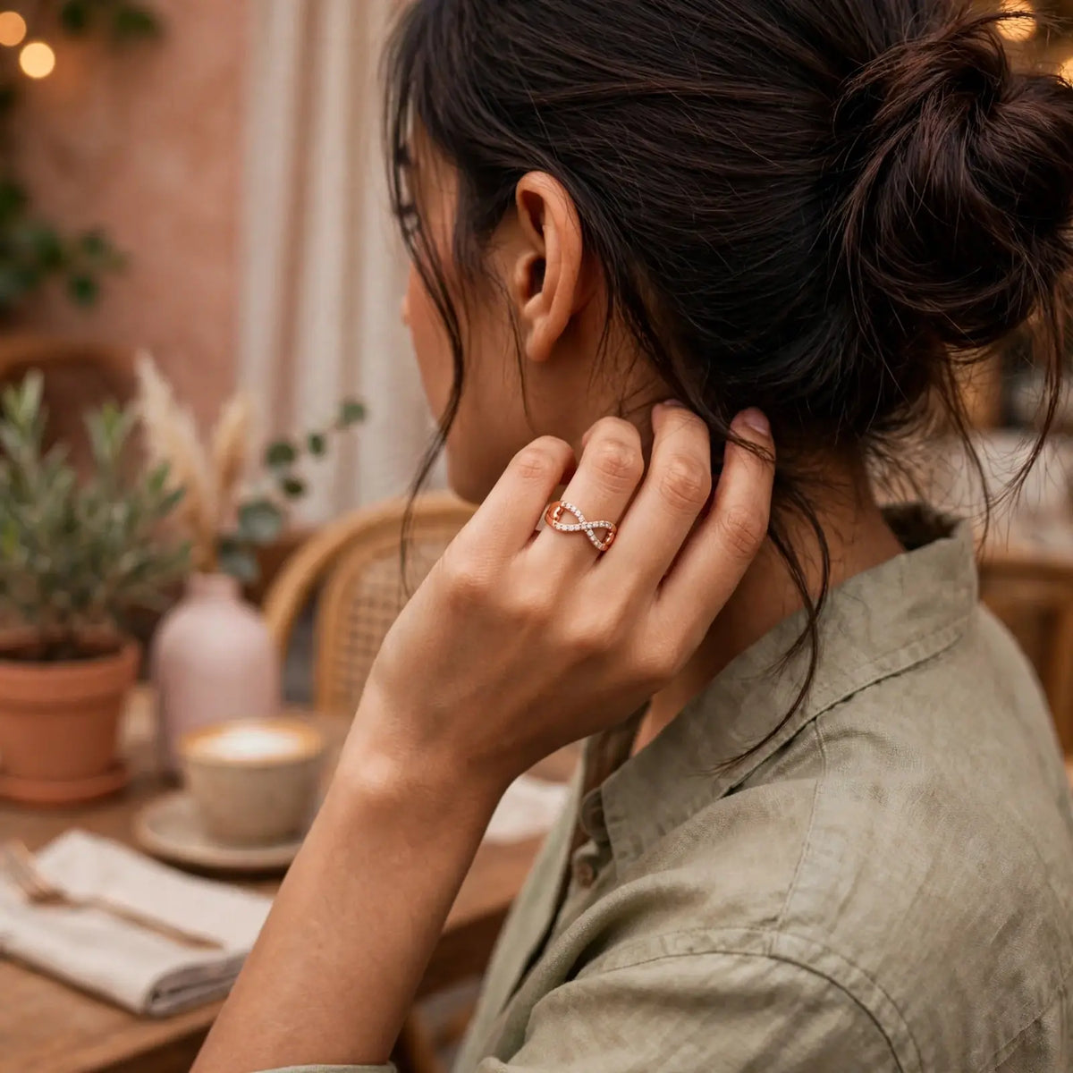 A woman in a cafe setting wearing a delicate infinity ring with a rose gold and white crystal design on her finger, seen from a side-back angle.