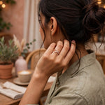 A woman in a cafe setting wearing a delicate infinity ring with a rose gold and white crystal design on her finger, seen from a side-back angle.
