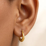 A close-up of a person's ear wearing a small textured gold hoop earring against a soft light background.
