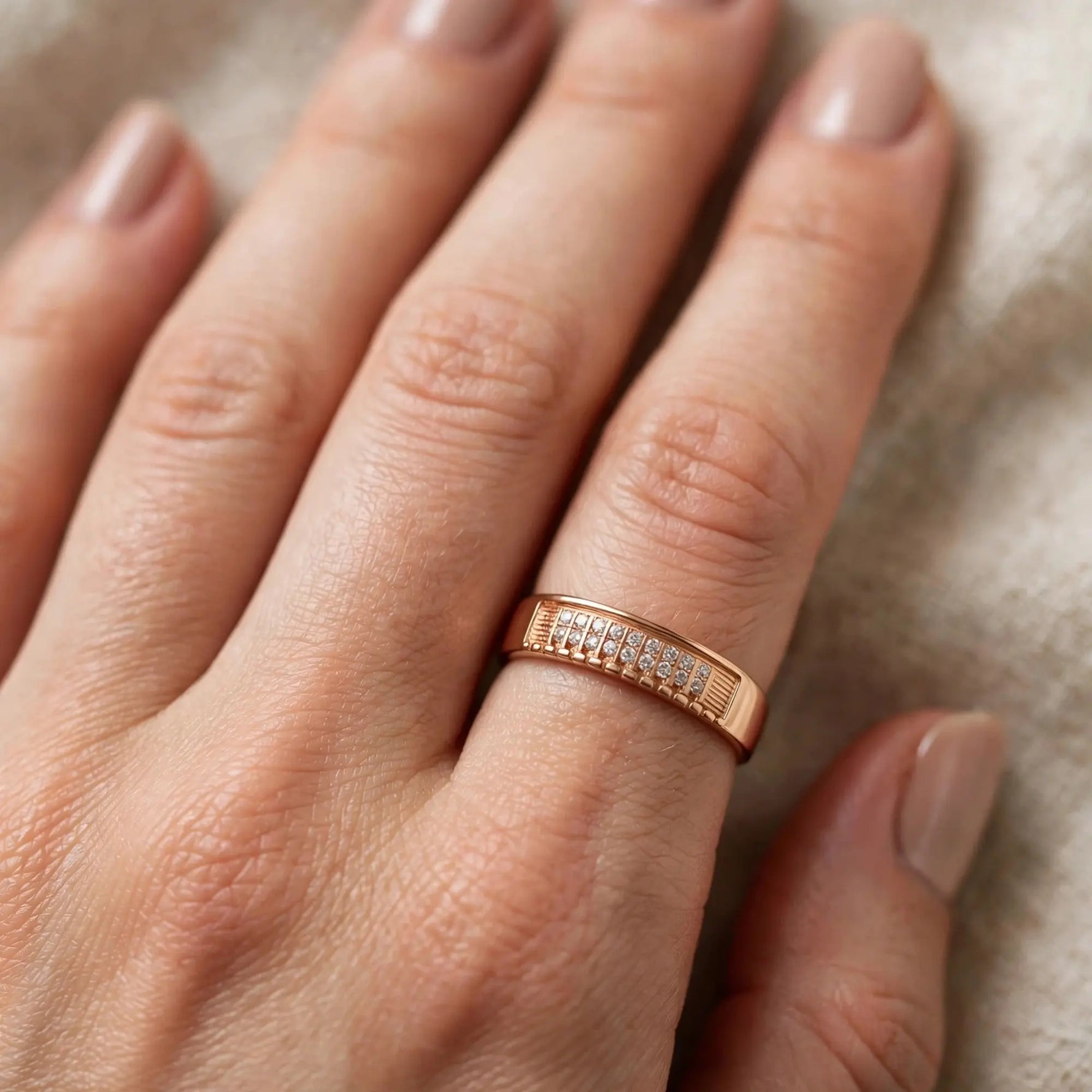A close-up of a hand wearing a slim rose gold band ring with a row of small clear stones on the side, set against a soft beige textured background.