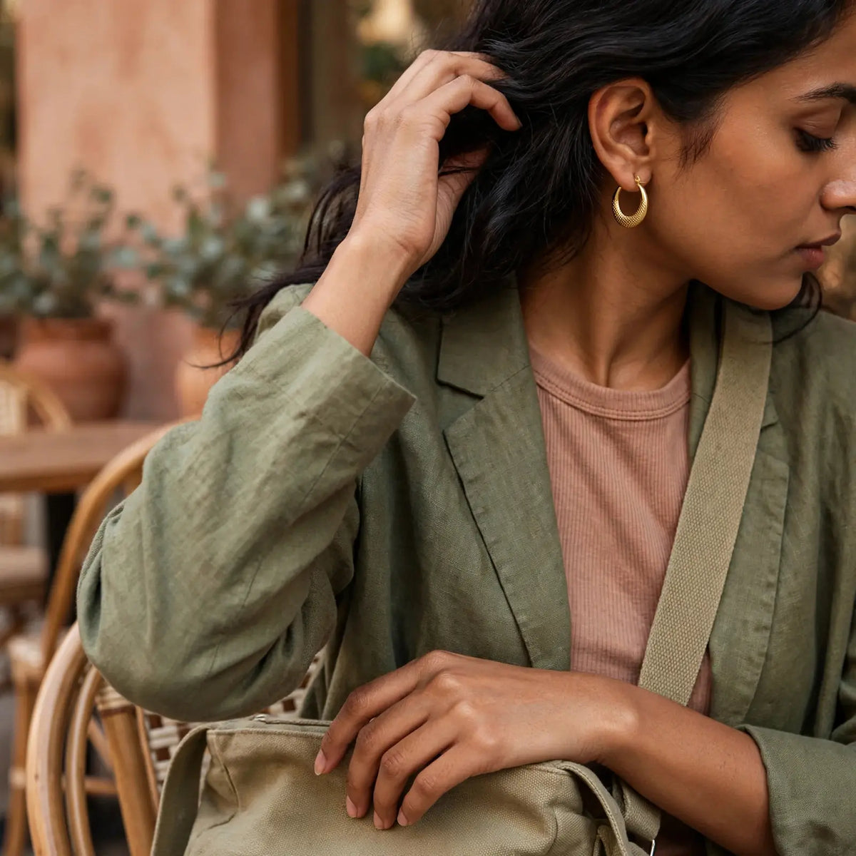 A woman wearing a muted olive green linen blazer over a dusty rose ribbed t-shirt, accessorized with a matching olive green shoulder bag and gold hoop earrings, in a warm, softly blurred cafe setting.
