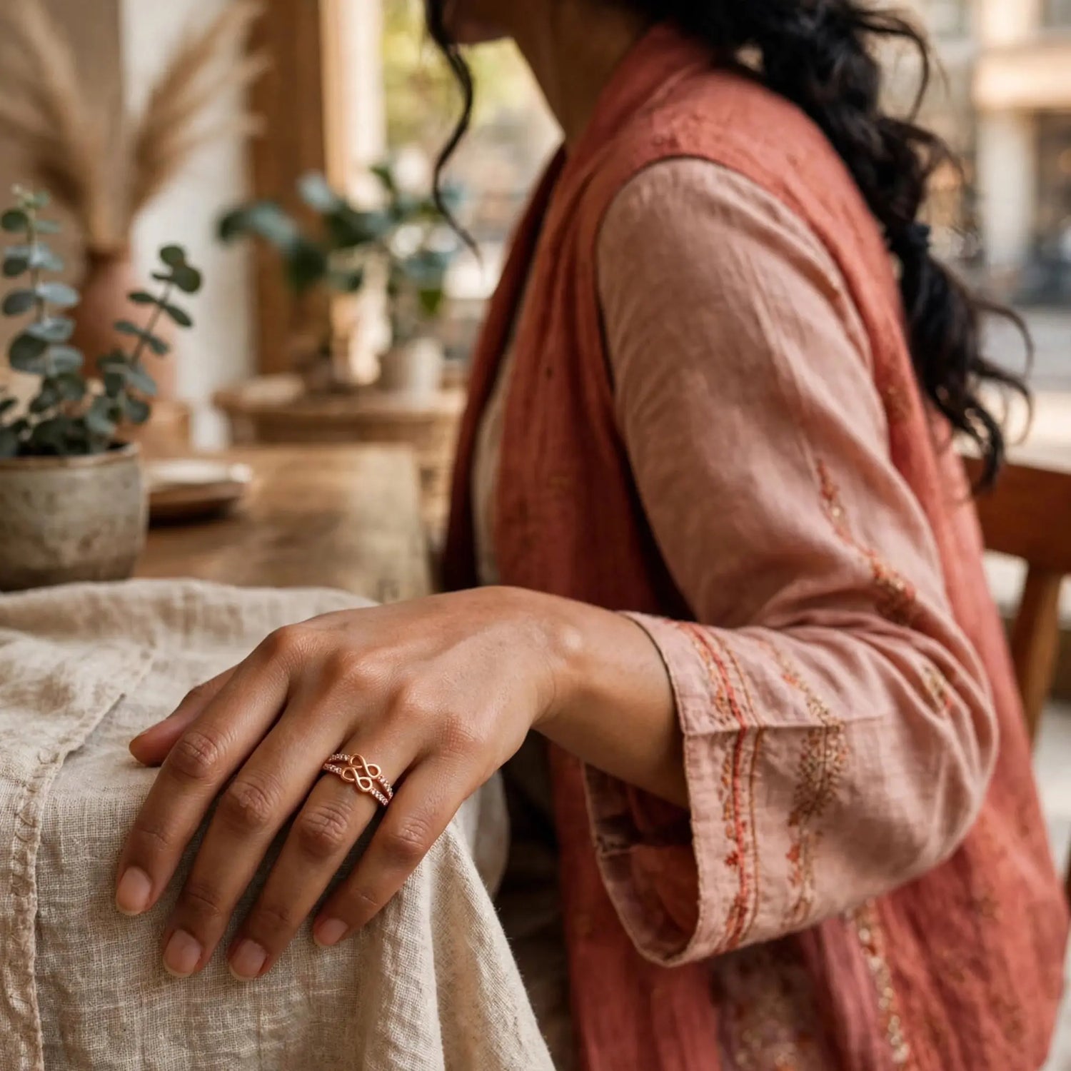 A close-up lifestyle shot of a woman wearing a delicate rose gold ring with an intricate knot design on her finger, dressed in a dusty pink embroidered vest over a beige linen shirt, with a warm bohemian background.