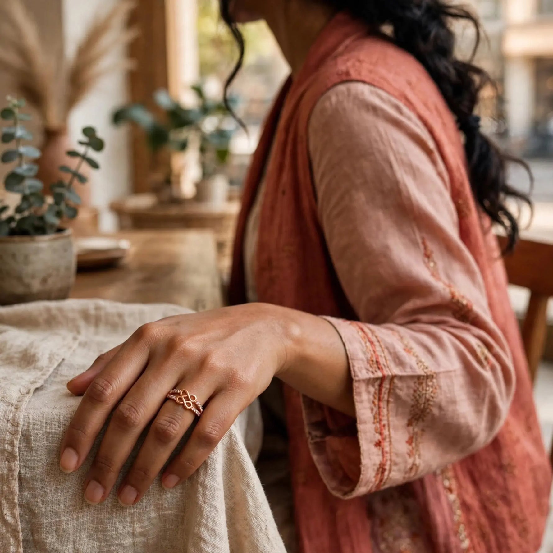 A close-up lifestyle shot of a woman wearing a delicate rose gold ring with an intricate knot design on her finger, dressed in a dusty pink embroidered vest over a beige linen shirt, with a warm bohemian background.