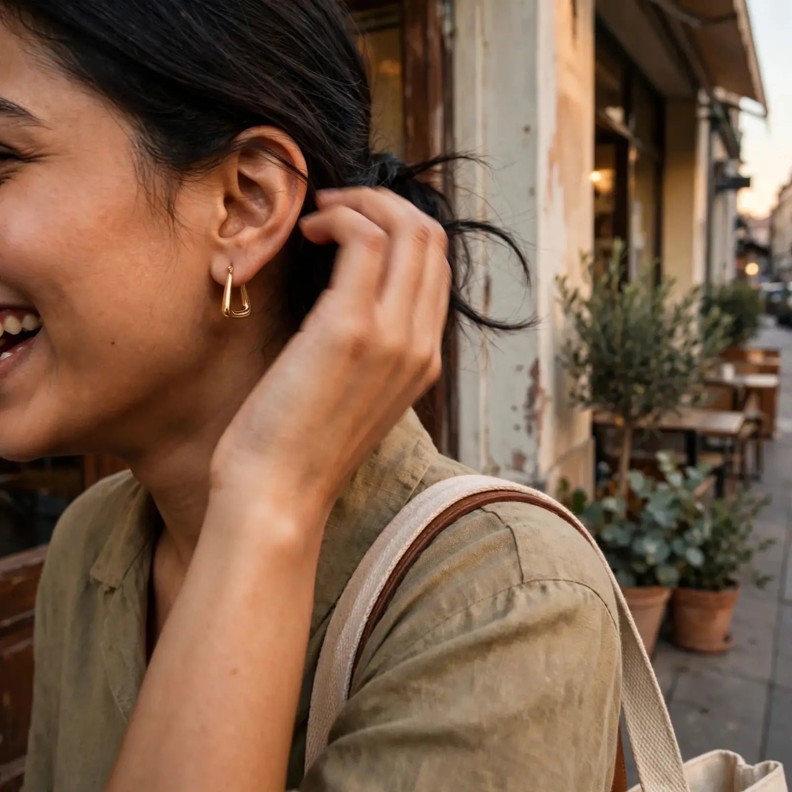 A woman outdoors wearing a small gold huggie hoop earring, with her hand near her hair and a beige shoulder bag visible.